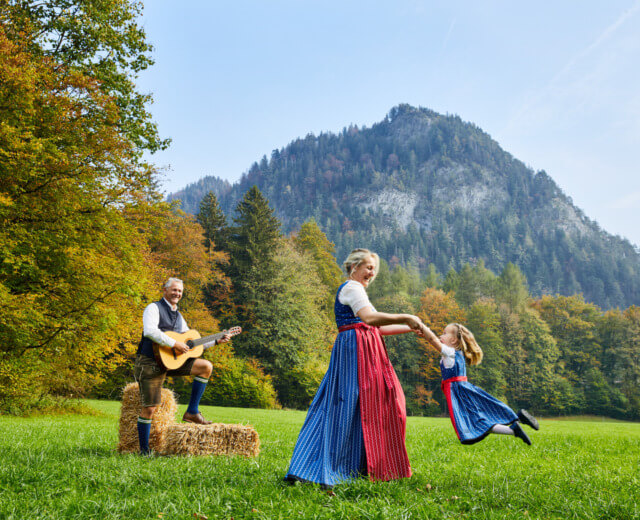 Herbstmusik Eine Frau wirbelt ein Mädchen in traditioneller Kleidung auf einer Wiese auf dem Biobauernhof herum, während in der Nähe Berge und Gitarrenmusik zu hören sind.