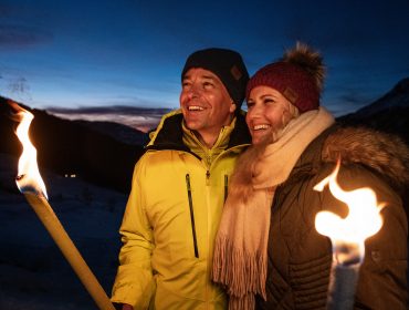 Ein lächelndes Paar in Winterjacken hält in der Abenddämmerung Fackeln in der Hand und stimmt sich inmitten verschneiter Berge auf den Advent ein.