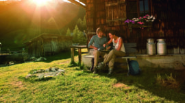 Pärchen vor Almhütte – Almsommer Hauptsujet 2012 Zwei Personen entspannen sich an einer Selbstversorgerhütte im Sonnenlicht mit Blick auf die Berge im Hintergrund.