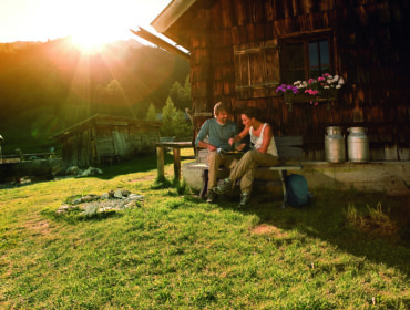 Zwei Personen entspannen sich an einer Selbstversorgerhütte im Sonnenlicht mit Blick auf die Berge im Hintergrund.