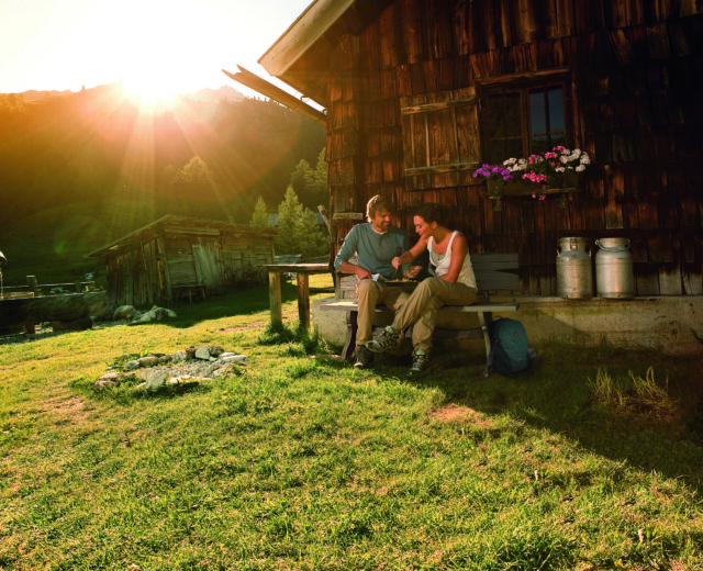 Pärchen vor Almhütte – Almsommer Hauptsujet 2012 Zwei Personen entspannen sich an einer Selbstversorgerhütte im Sonnenlicht mit Blick auf die Berge im Hintergrund.