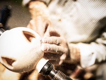 Nahaufnahme von Händen, die beim Bauernherbst Holz auf einer Drehbank bearbeiten, wobei Holzspäne fliegen.