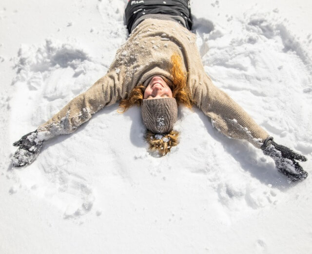 Schneeengel Person in Winterkleidung, die mit ausgestreckten Armen im Schnee am Wolfgangsee liegt, einen Schneeengel macht und lächelt.