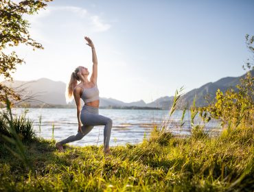 Frau übt Yoga im Freien an einem See in der alpinen Gesundheitsregion, umgeben von Gras und Bergen.