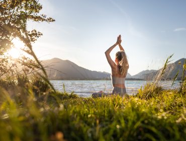 Frau übt Yoga an einem See bei Sonnenaufgang in der alpinen Gesundheitsregion, Berge im Hintergrund.