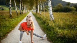 Eine Frau in Sportkleidung dehnt sich auf einem von Bäumen gesäumten Weg in der malerischen Hügellandschaft der alpinen Gesundheitsregion.