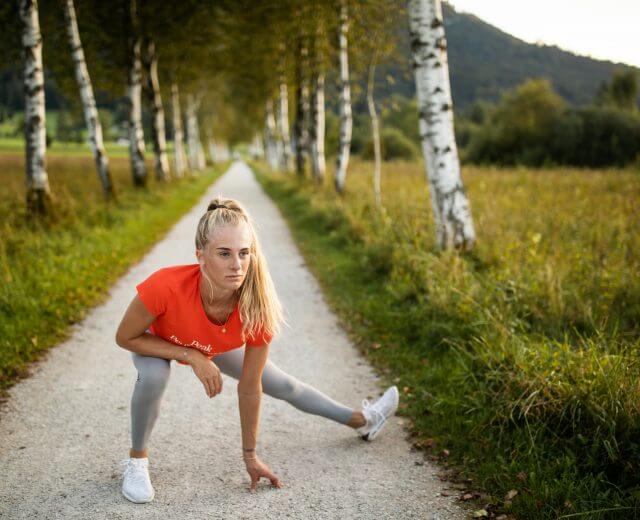 Eine Frau in Sportkleidung dehnt sich auf einem von Bäumen gesäumten Weg in der malerischen Hügellandschaft der alpinen Gesundheitsregion.