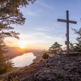 Holzkreuz auf einem Berggipfel mit Blick auf den Fuschlsee und Berge bei Sonnenuntergang, umgeben von Bäumen.