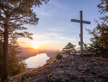 Holzkreuz auf einem Berggipfel mit Blick auf den Fuschlsee und Berge bei Sonnenuntergang, umgeben von Bäumen.