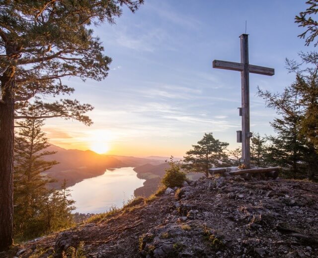 Holzkreuz auf einem Berggipfel mit Blick auf den Fuschlsee und Berge bei Sonnenuntergang, umgeben von Bäumen.
