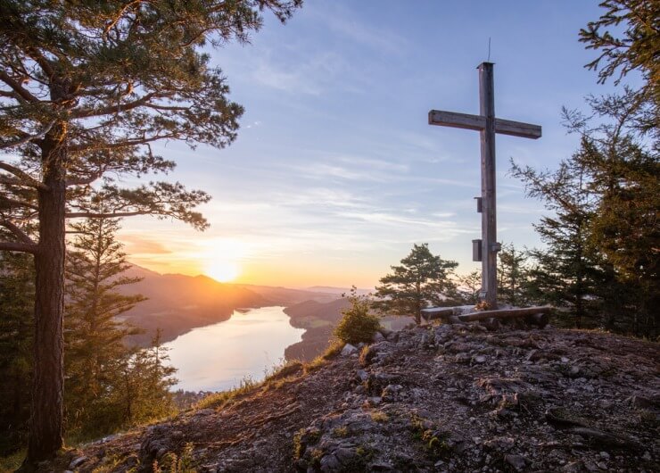 Holzkreuz auf einem Berggipfel mit Blick auf den Fuschlsee und Berge bei Sonnenuntergang, umgeben von Bäumen.