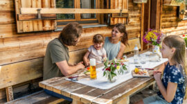 Eine vierköpfige Familie sitzt lächelnd an einem Picknicktisch vor einer rustikalen, blumengeschmückten, bewirtschafteten Hütte.
