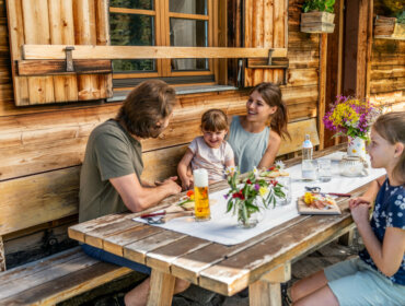 Eine vierköpfige Familie sitzt lächelnd an einem Picknicktisch vor einer rustikalen, blumengeschmückten, bewirtschafteten Hütte.
