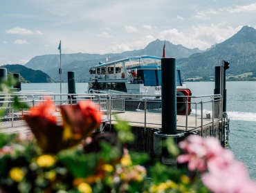 Ein Boot, das an einer Anlegestelle am Wolfgangsee angelegt hat, mit Bergen im Hintergrund und Blumen im Vordergrund.