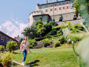 Burg Hohenwerfen Ein Ehepaar spaziert durch einen sonnigen Garten mit einem historischen österreichischen Schloss - entdecken Sie das alles mit der SalzburgerLand Card.