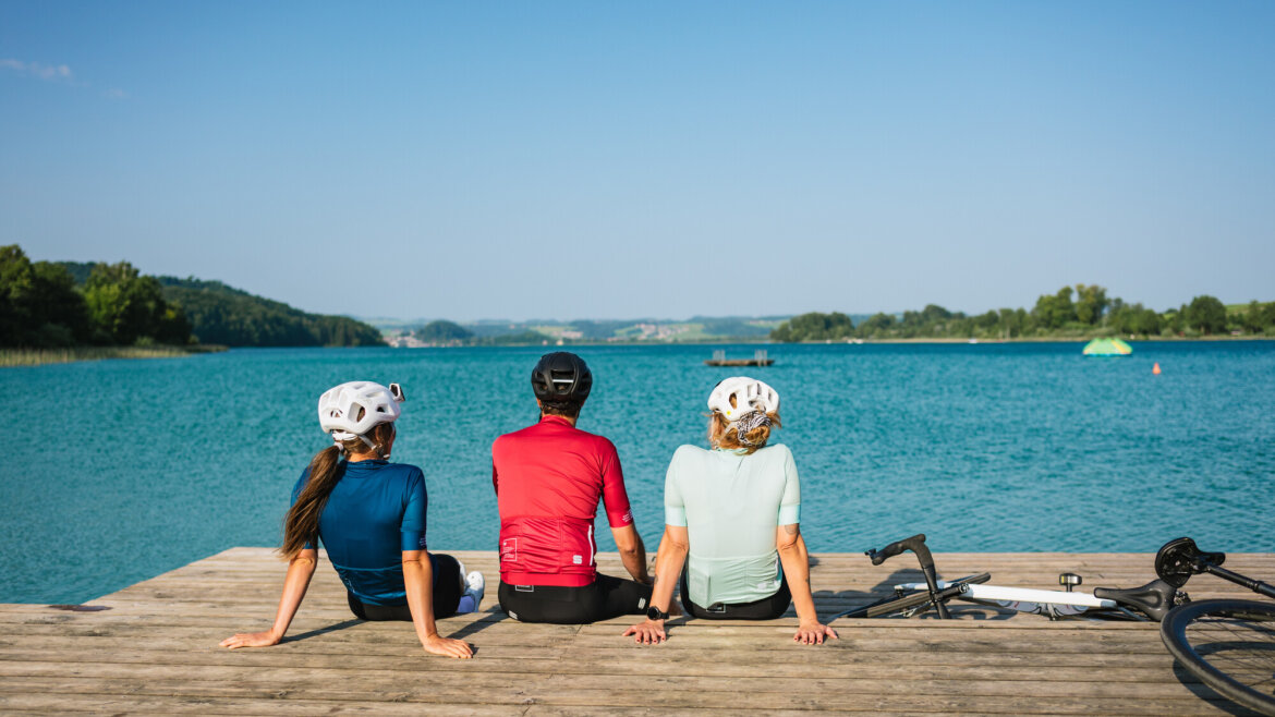 Drei Rennradfahrer mit Helmen entspannen sich an einem sonnigen Tag auf einem Steg an einem blauen See, Fahrräder neben ihnen.