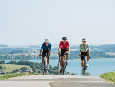 Drei Radfahrer genießen das Radfahren auf einer Landstraße mit einem See und einer malerischen Landschaft im Hintergrund.