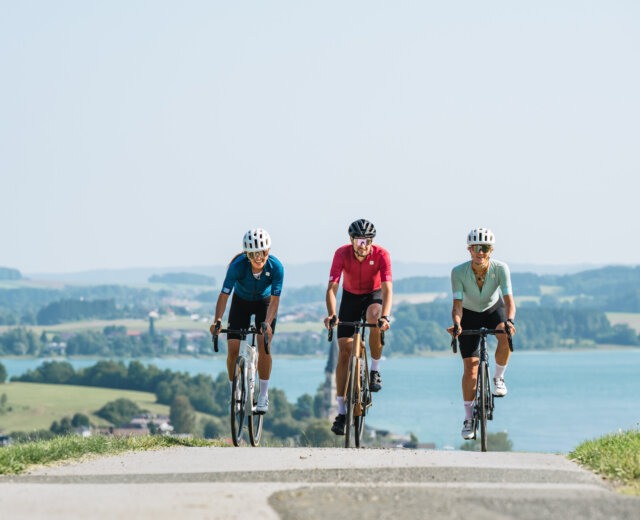 Rennrad fahren im Salzburger Seenland Drei Radfahrer genießen das Radfahren auf einer Landstraße mit einem See und einer malerischen Landschaft im Hintergrund.