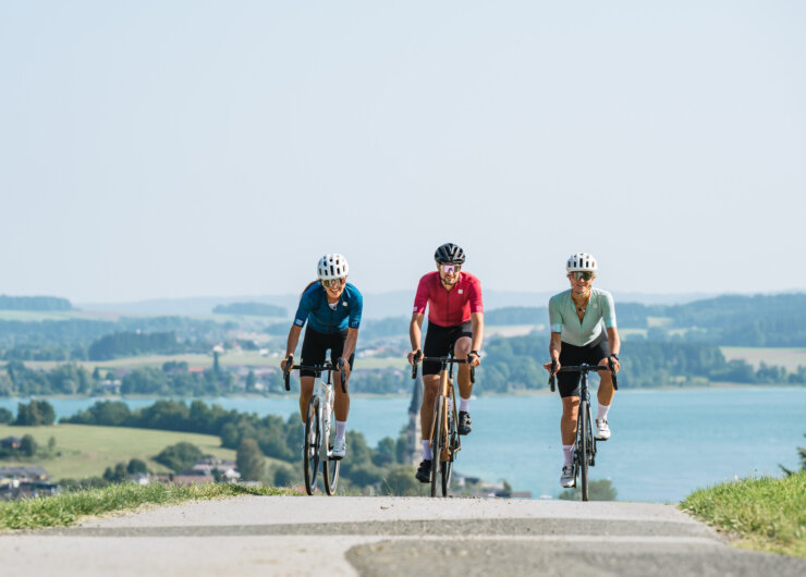 Drei Radfahrer genießen das Radfahren auf einer Landstraße mit einem See und einer malerischen Landschaft im Hintergrund.