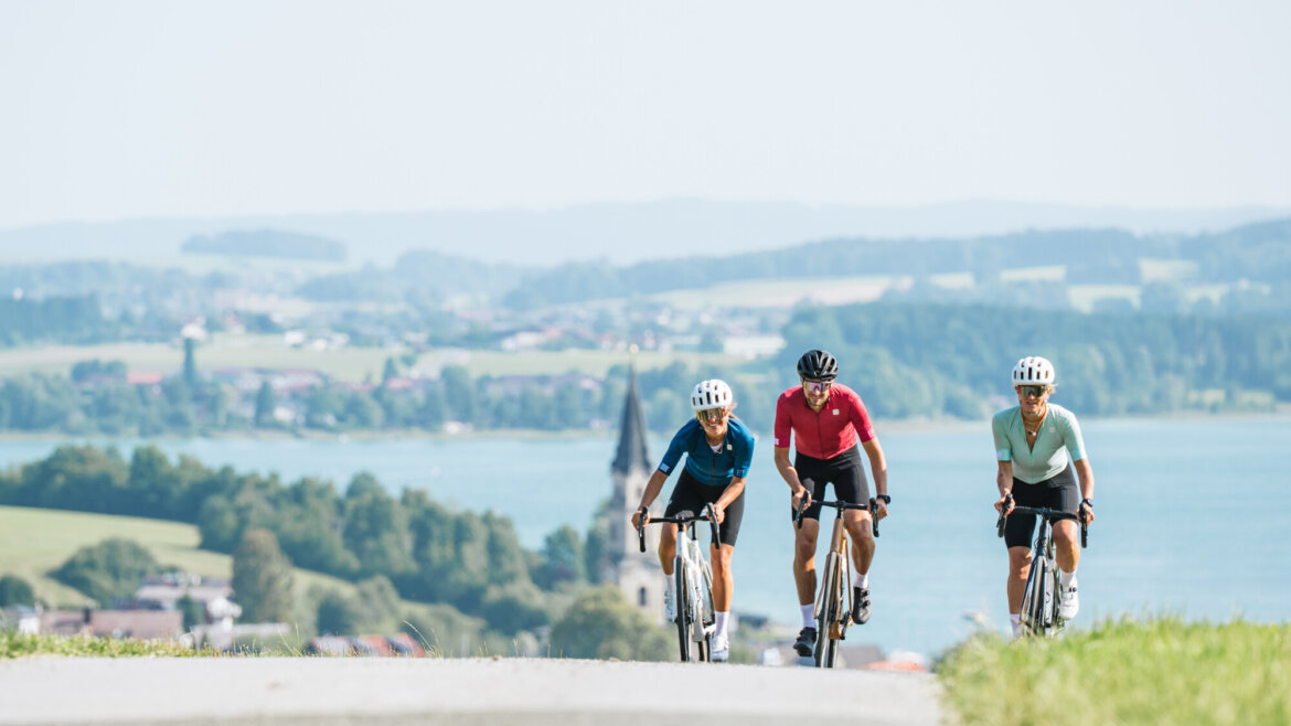 Drei Radfahrer genießen das Rennradfahren bergauf auf einer Landstraße mit einem malerischen See und einem Dorf dahinter.