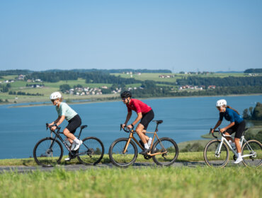 Drei Radfahrer genießen das Rennradfahren auf einem Seeuferweg mit grünen Hügeln und einem Dorf im Hintergrund.