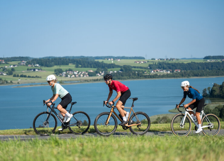 Drei Radfahrer genießen das Rennradfahren auf einem Seeuferweg mit grünen Hügeln und einem Dorf im Hintergrund.
