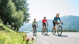 Drei Radfahrer genießen einen sonnigen Radmarathon in der Nähe von Bergen und Bäumen, im Vordergrund blühen Wildblumen.