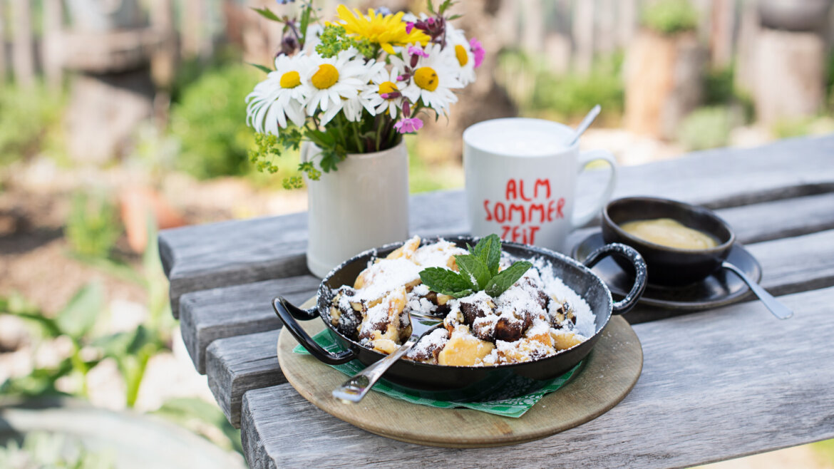 Eine Pfanne mit Via Culinaria-Dessert, bestreut mit Puderzucker, einer Blumenvase und einem Becher auf einem Holztisch. (vergrößerte Ansicht)
