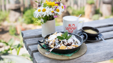 Eine Pfanne mit Via Culinaria-Dessert, bestreut mit Puderzucker, einer Blumenvase und einem Becher auf einem Holztisch.
