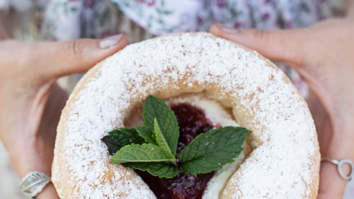 Eine Person hält ein Via Culinaria-Gebäck mit Puderzucker, Marmelade und Minzblättern in der Hand und trägt eine geblümte Schürze.