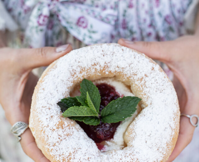 Eine Person hält ein Via Culinaria-Gebäck mit Puderzucker, Marmelade und Minzblättern in der Hand und trägt eine geblümte Schürze.