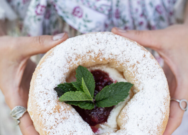Eine Person hält ein Via Culinaria-Gebäck mit Puderzucker, Marmelade und Minzblättern in der Hand und trägt eine geblümte Schürze.