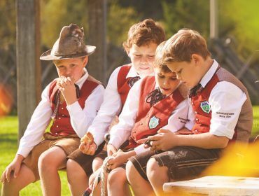 Vier Jungen in österreichischem Outfit sitzen auf einer Bank beim Bauernherbst; zwei schauen etwas auf einem Telefon an.