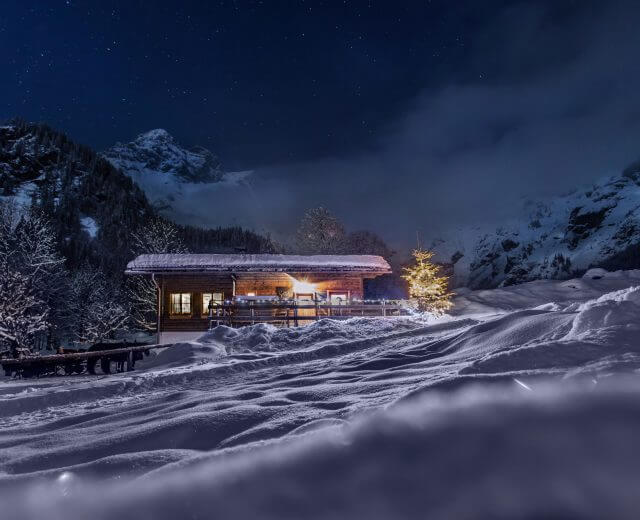 Hütte Adventzeit Eine gemütliche Adventshütte erstrahlt in warmen Lichtern inmitten verschneiter Berge und Bäume unter einem sternenklaren Nachthimmel.