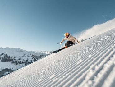 Ein Skifahrer mit Helm fährt unter freiem Himmel eine präparierte Schneepiste mit Bergen im Hintergrund hinunter.