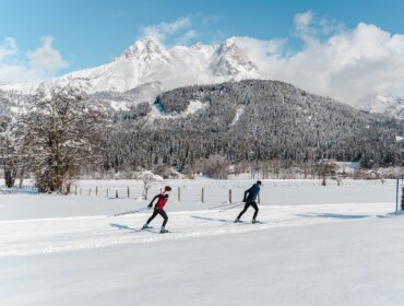 Zwei Personen beim Skilanglauf in der österreichischen Region Saalfelden, mit verschneiten Bergen und Bäumen im Hintergrund.