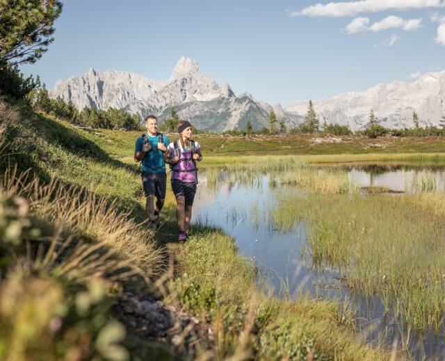 Zwei Personen beim Trailrunning an einem Bergsee in der Salzburger Sportwelt, umgeben von saftigem Gras und felsigen Gipfeln.