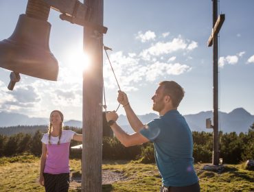 Ein Mann läutet eine große Glocke im Freien, während Eben in der Nähe lächelt, mit Bergen und Sonne im Hintergrund.