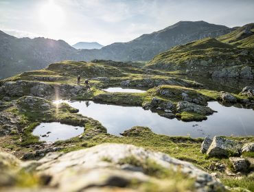 Zwei Personen fahren Mountainbike und wandern in der Nachmittagssonne in der Nähe kleiner Teiche auf einem grasbewachsenen, felsigen Berg.