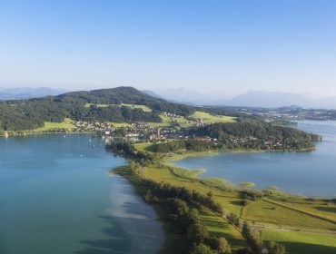 Luftaufnahme einer grünen Halbinsel im Salzburger Seenland, blaues Wasser und ein kleines Dorf unter einem klaren Himmel.