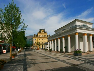 Eine breite Fußgängerzone in Bad Ischl mit Bäumen, weißen Säulen und einem gelben historischen Gebäude unter blauem Himmel.