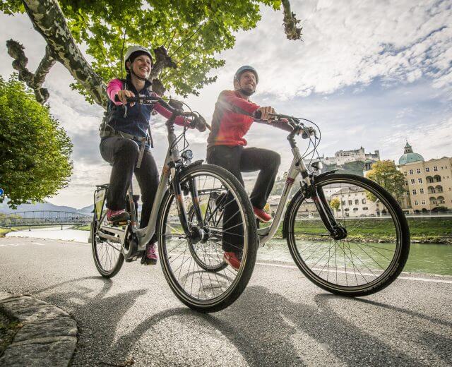 Zwei Radfahrer fahren am Ufer der Salzach auf dem Tauernradweg durch die Stadt Salzburg.