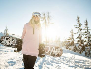 Ein lächelnder Snowboarder in einem rosa Kapuzenpulli hält ein Snowboard auf einem sonnigen, verschneiten Berg mit Bäumen im Hintergrund.