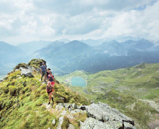Drei Wanderer wandern auf einem grasbewachsenen Bergrücken in der Nähe von Obertauern, mit einem malerischen See und Gipfeln im Hintergrund.