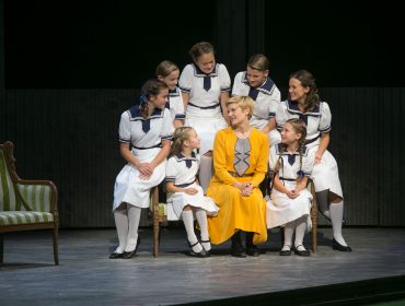 Eine Frau in Gelb sitzt mit Mädchen in Matrosenkleidern auf der Bühne und lächelt nach einer Sound of Music Tour.