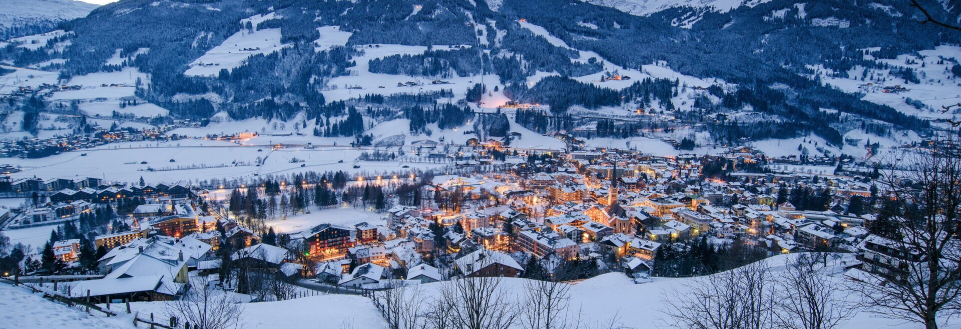 Verschneites Dorf Bad Hofgastein mit leuchtenden Lichtern, eingebettet in ein Tal, Berge in der Abenddämmerung unter einem bewölkten Himmel.