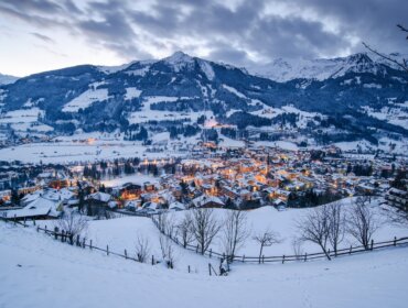 Bad Hofgastein Verschneites Dorf Bad Hofgastein mit leuchtenden Lichtern, eingebettet in ein Tal, Berge in der Abenddämmerung unter einem bewölkten Himmel.