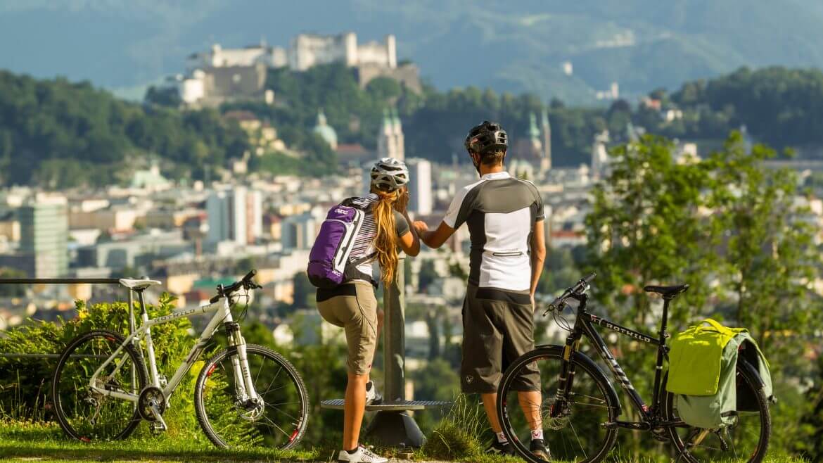 Ein Pärchen steht neben den Fahrrädern und genießt den imposanten Blick auf die Stadt Salzburg, einer der Höhepunkte am Tauernradweg.
