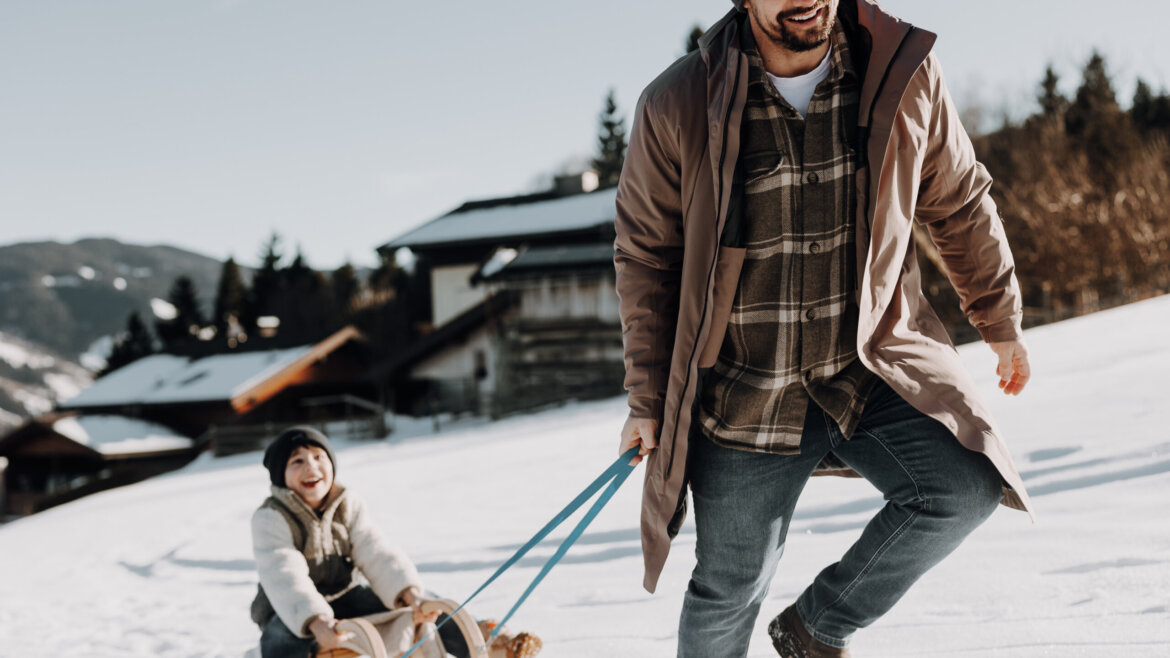 © Tourismusverband Bischofshofen | two minds Studio Ein lächelnder Mann zieht ein Kind auf einem Schlitten durch das verschneite Bischofshofen in der Nähe von Holzhütten und Bäumen. (vergrößerte Ansicht)