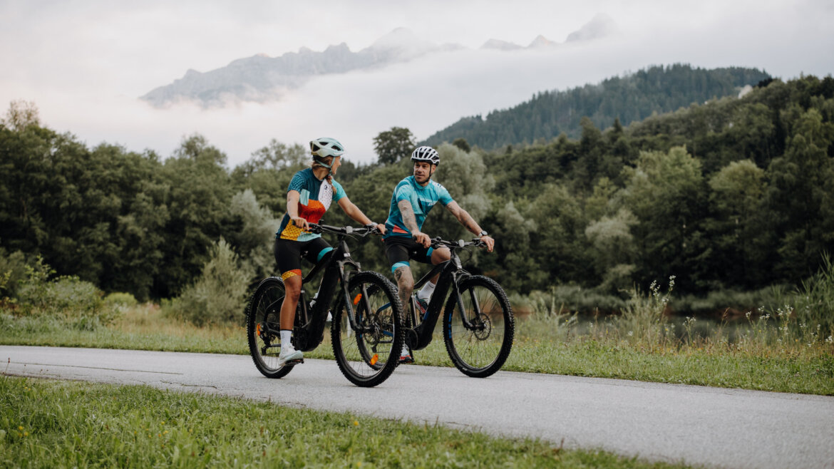 © Tourismusverband Bischofshofen | two minds Studio Zwei Radfahrer fahren auf einem asphaltierten Weg in der Nähe von Bischofshofen, umgeben von üppigem Grün und nebligen Bergen. (vergrößerte Ansicht)